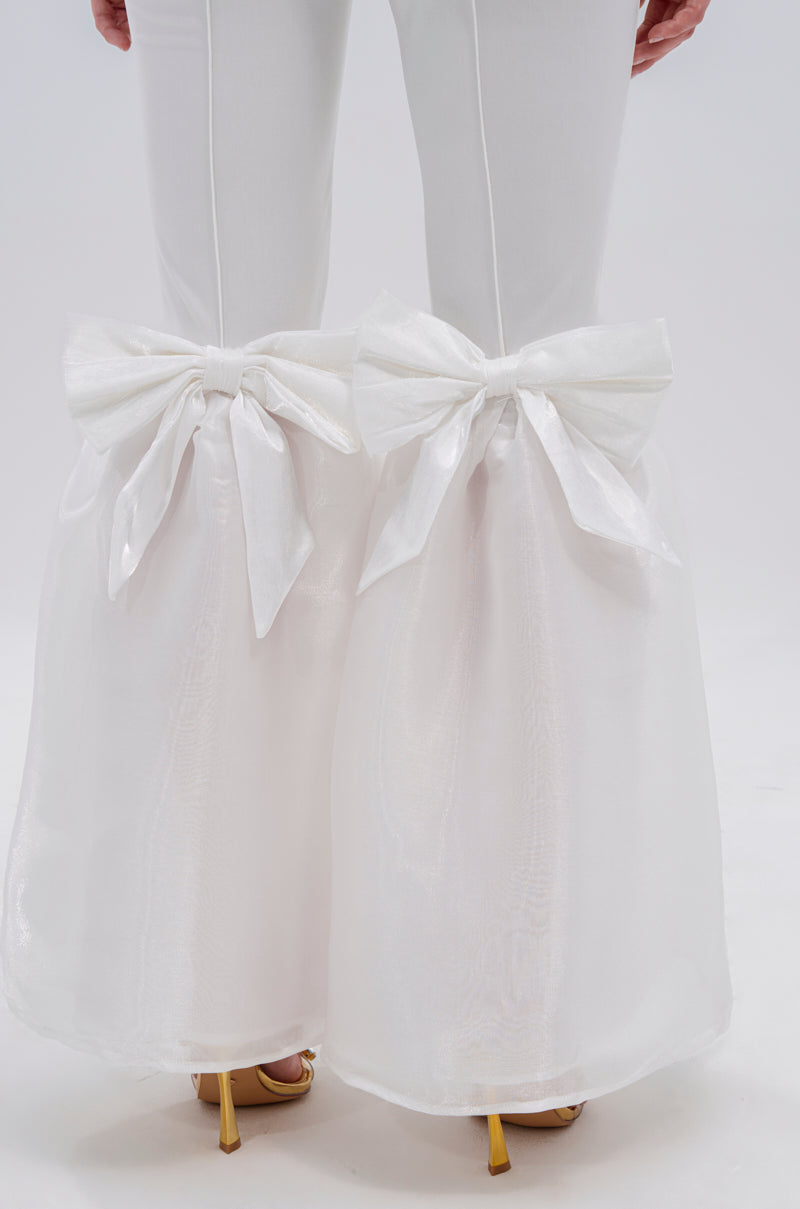 Close-up of lower legs in the LILY TROUSER WITH BOW, showcasing high-rise white flared pants with sheer fabric over the flare and large satin bows at each knee, styled with gold high-heeled shoes for elegance.