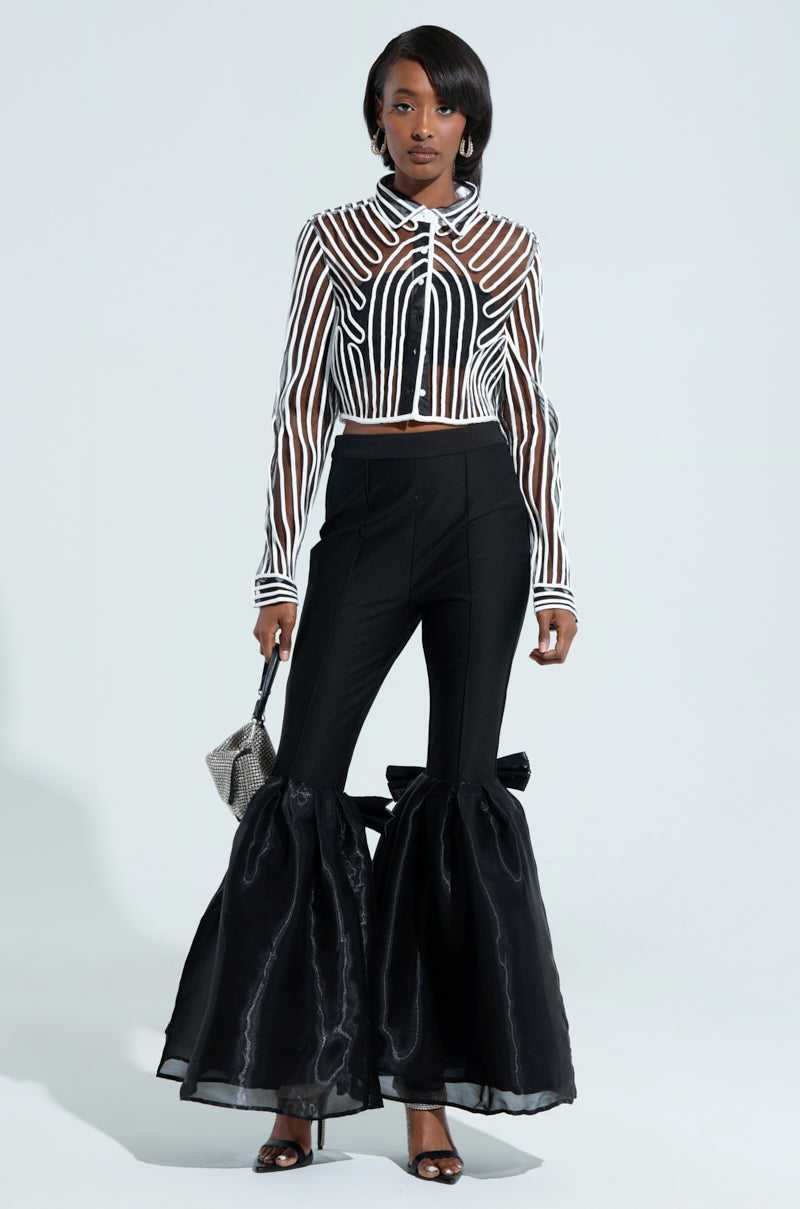 A woman wears a chic sheer black-and-white striped cropped top and the LILY TROUSER WITH BOW IN BLACK, known for its high-rise fit and dramatic flared bottoms. She accessorizes with a small silver handbag and black heels.