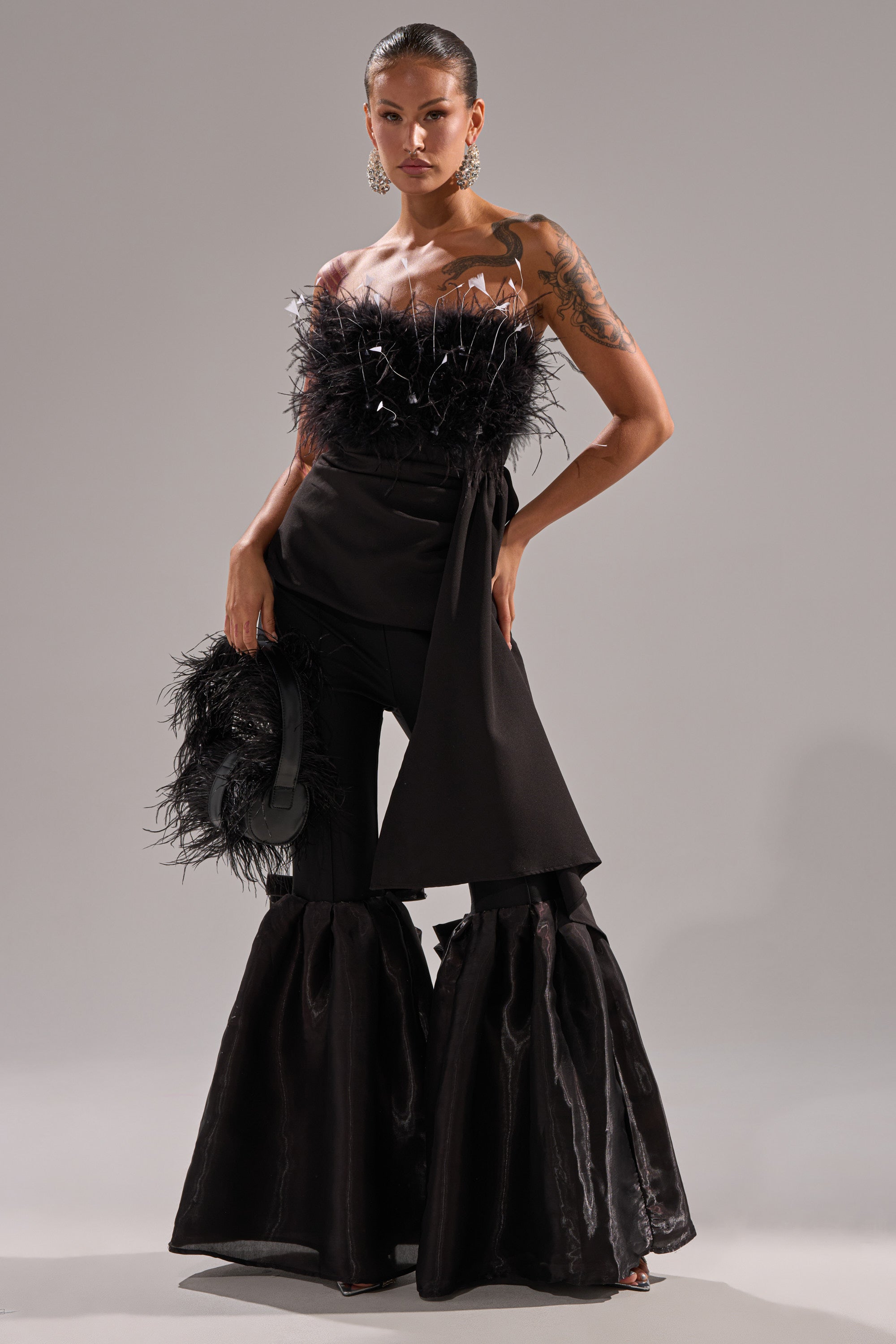 A woman poses in a dramatic black outfit featuring the LILY TROUSER WITH BOW IN BLACK, styled with a feathered strapless top, bold earrings, and a matching feathered accessory. She showcases a tattoo on her arm.
