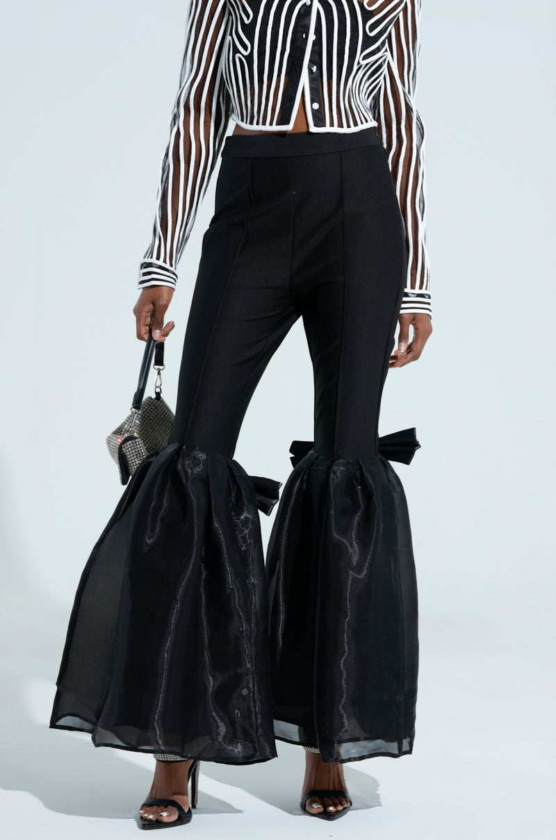 Wearing the LILY TROUSER WITH BOW IN BLACK, featuring a high-rise fit, sheer voluminous flared legs, and satin knee bows, paired with a black-and-white striped sheer top, heeled sandals, and a small handbag.