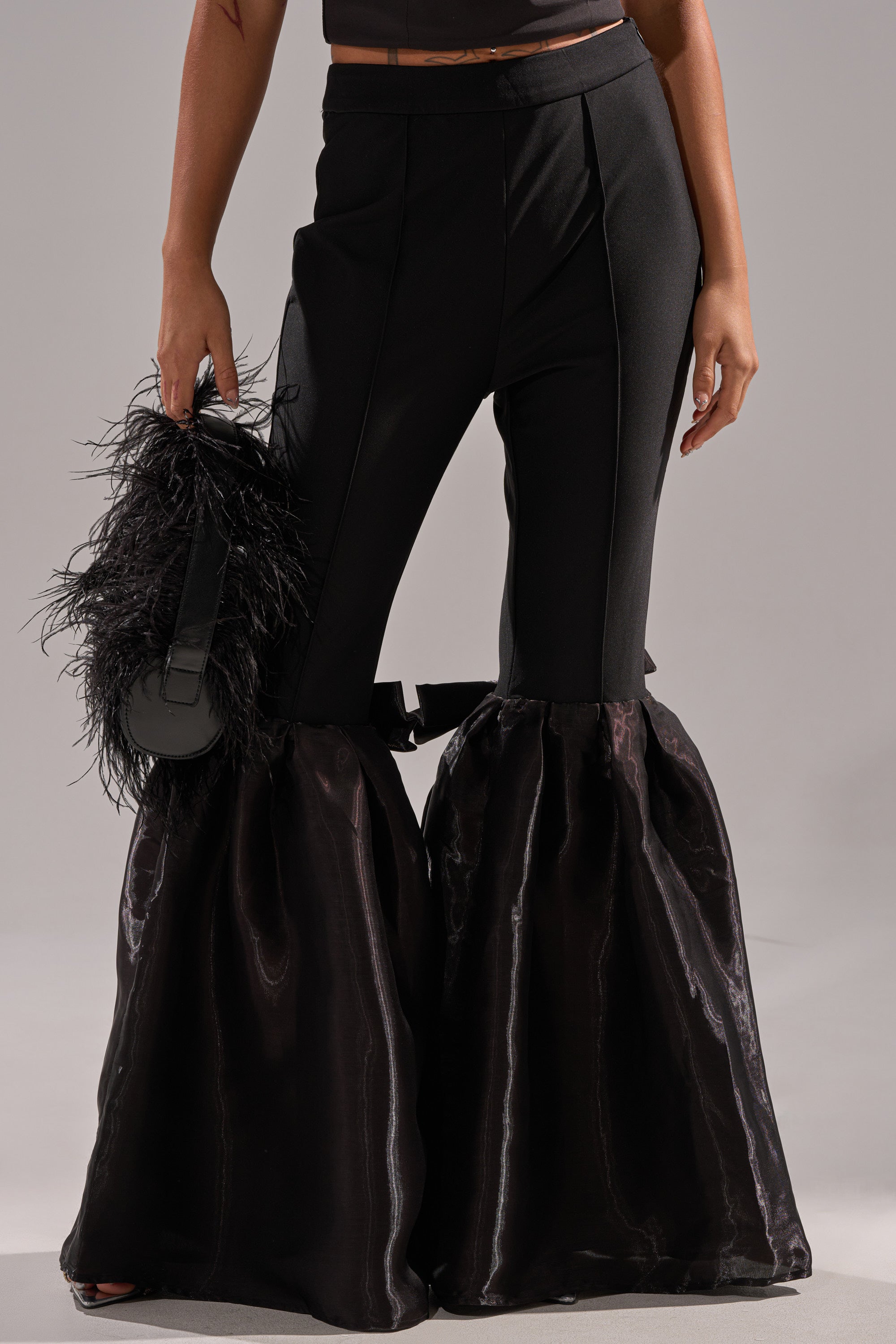 Wearing the LILY TROUSER WITH BOW IN BLACK, which features high-rise, flared legs with voluminous fabric, and holding a black feathered clutch against a plain background.