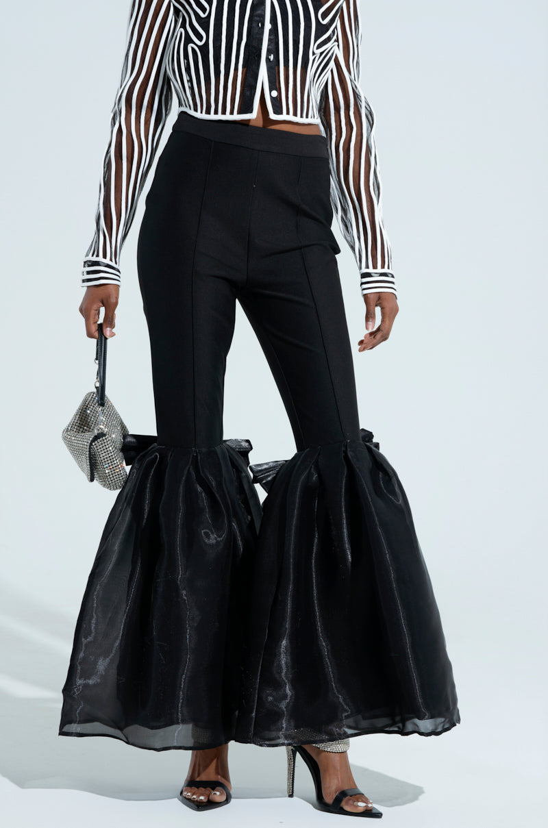A person wearing LILY TROUSER WITH BOW IN BLACK high-waisted pants with voluminous ruffled hems, a sheer striped black and white cropped jacket, satin bows, black heels, and carrying a small silver embellished handbag.