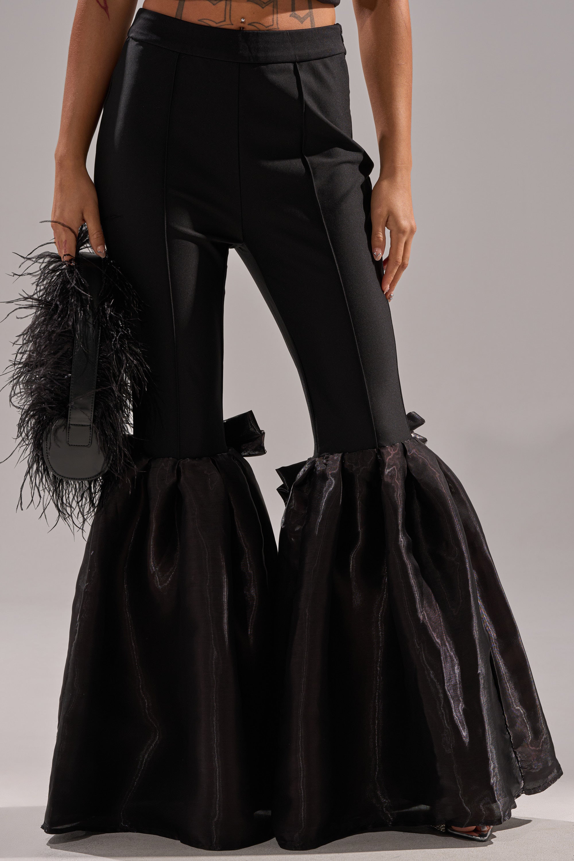 A person wears the LILY TROUSER WITH BOW IN BLACK, featuring high-rise flare legs with dramatic organza flares at the calves, and holds a black feather-detailed bag against a plain gray background.