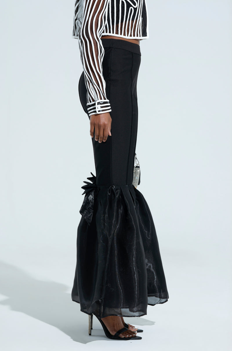 A person wears black LILY TROUSER WITH BOW IN BLACK, featuring sheer voluminous ruffle cuffs and a high-rise fit ‘n flare style, paired with a black and white striped sheer top and black high-heeled sandals, standing against a plain background.
