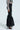 A person is elegantly dressed in an AKIRA Label ensemble, featuring a black and white striped sheer long-sleeve top, paired effortlessly with Lily Trouser With Bow In Black, high heels, and a small clutch bag against a plain background.