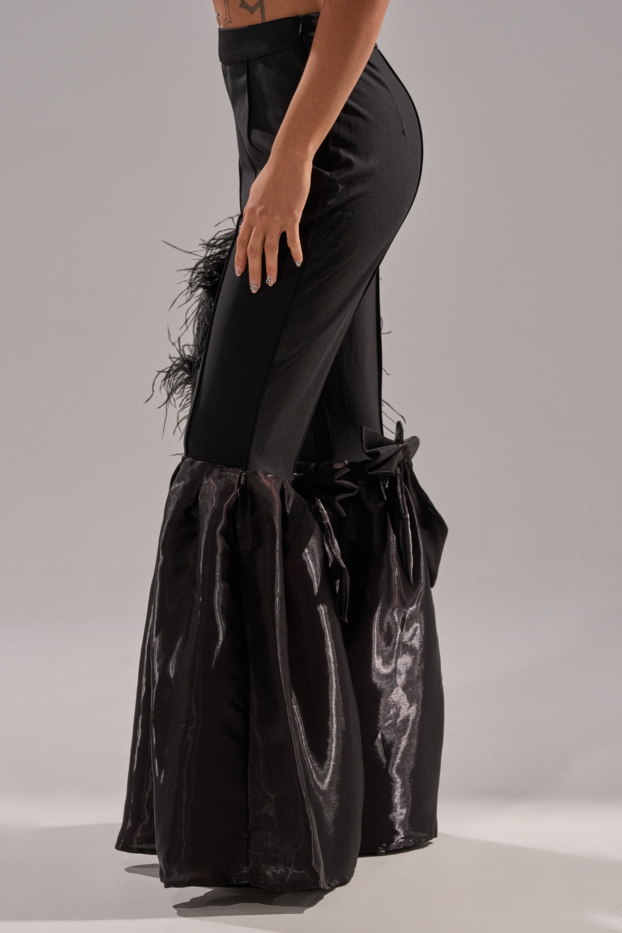 A person models the LILY TROUSER WITH BOW IN BLACK: high-rise, fitted through the hip and thigh with dramatic, shiny flared legs and bow details. The photo features a plain light background.