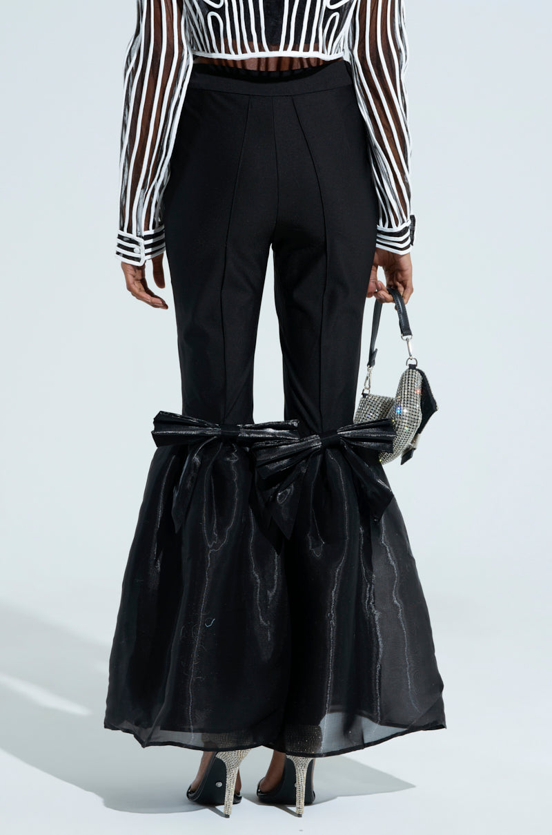 A person wearing LILY TROUSER WITH BOW IN BLACK, featuring a high-rise fit and flare design with satin bows, paired with a sheer black-and-white striped top, small silver purse, and pointed heels.