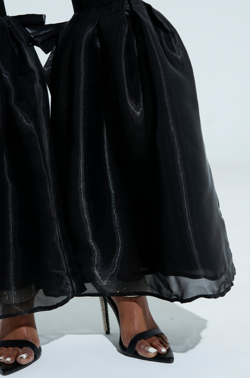A close-up shows only the lower legs and feet of a person wearing the LILY TROUSER WITH BOW IN BLACK, paired with open-toe black ankle-strap heels and white-painted toenails, set against a plain background.