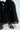 Wearing the elegant AKIRA Label LILY TROUSER WITH BOW IN BLACK, a person stands against a plain white background in open-toed black high heels. The tulle pants have a subtle sheen, adding sophistication to the look.