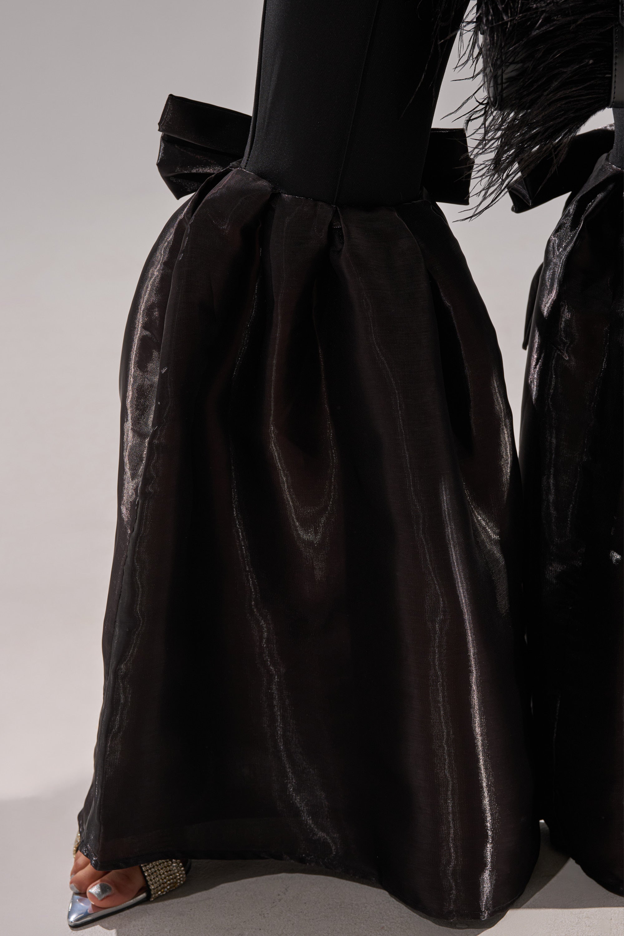 Wearing the LILY TROUSER WITH BOW IN BLACK—shiny, high-rise fit and flare pants with a large satin waist bow—paired with black feathered sleeve accents and silver open-toe shoes featuring sparkling embellishments.