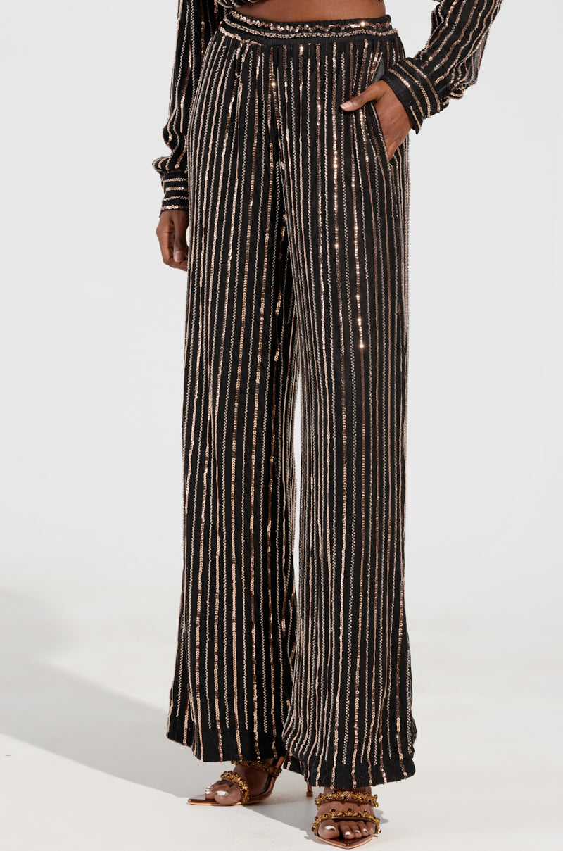 A person is dressed in the Flashing Lights Pant, which are black wide-leg pants adorned with vertical sequin embellishments. They pair it with a matching long-sleeved top featuring the same pattern. This holiday-ready ensemble is completed with gold sandals against a plain, light-colored background.