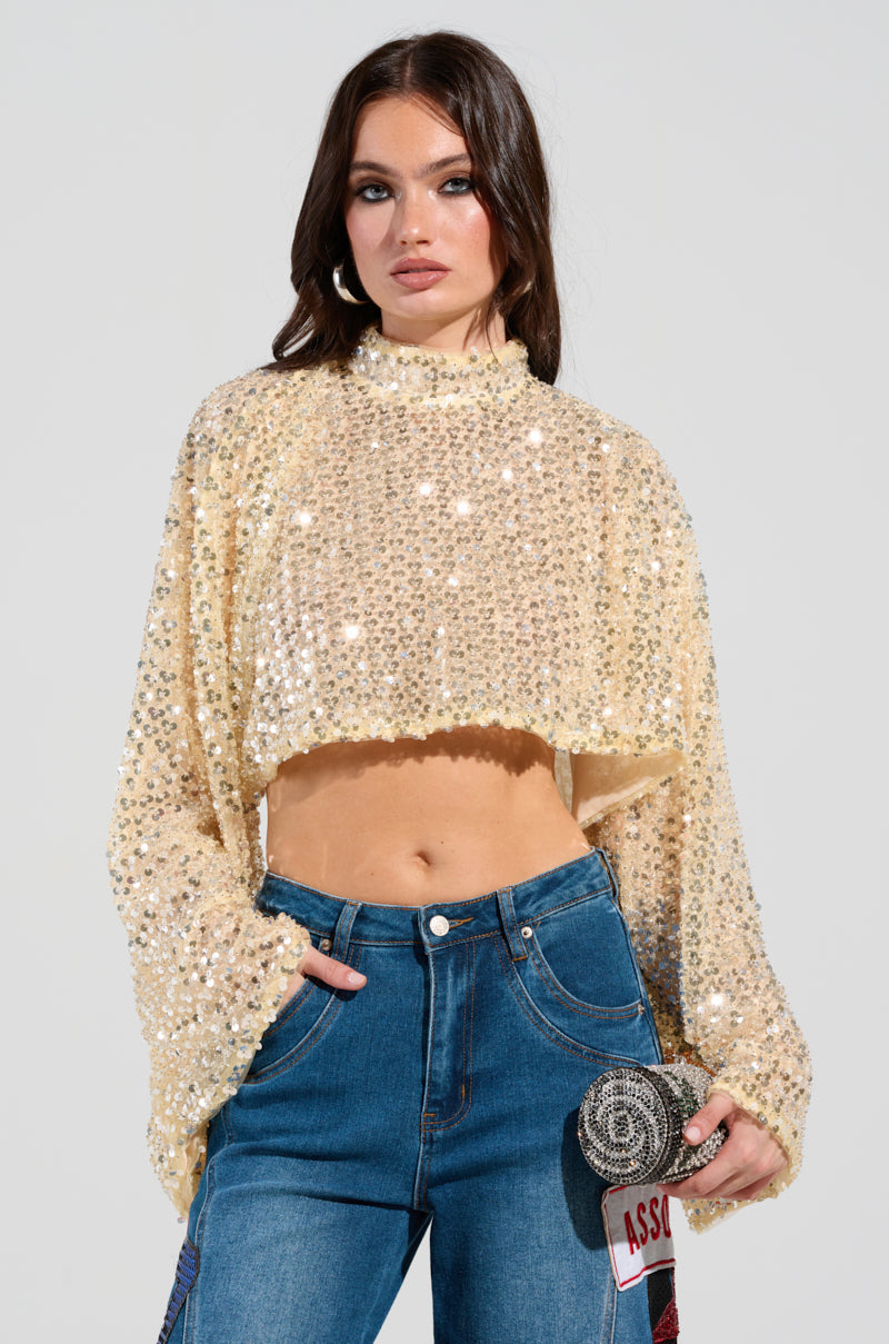 A woman poses confidently in the SLYLINE SEQUIN BLOUSE, a glittery beige top featuring wide sleeves, paired with high-waisted blue jeans. She holds a small, round sequined clutch that complements her holiday party look and stands against a plain background.