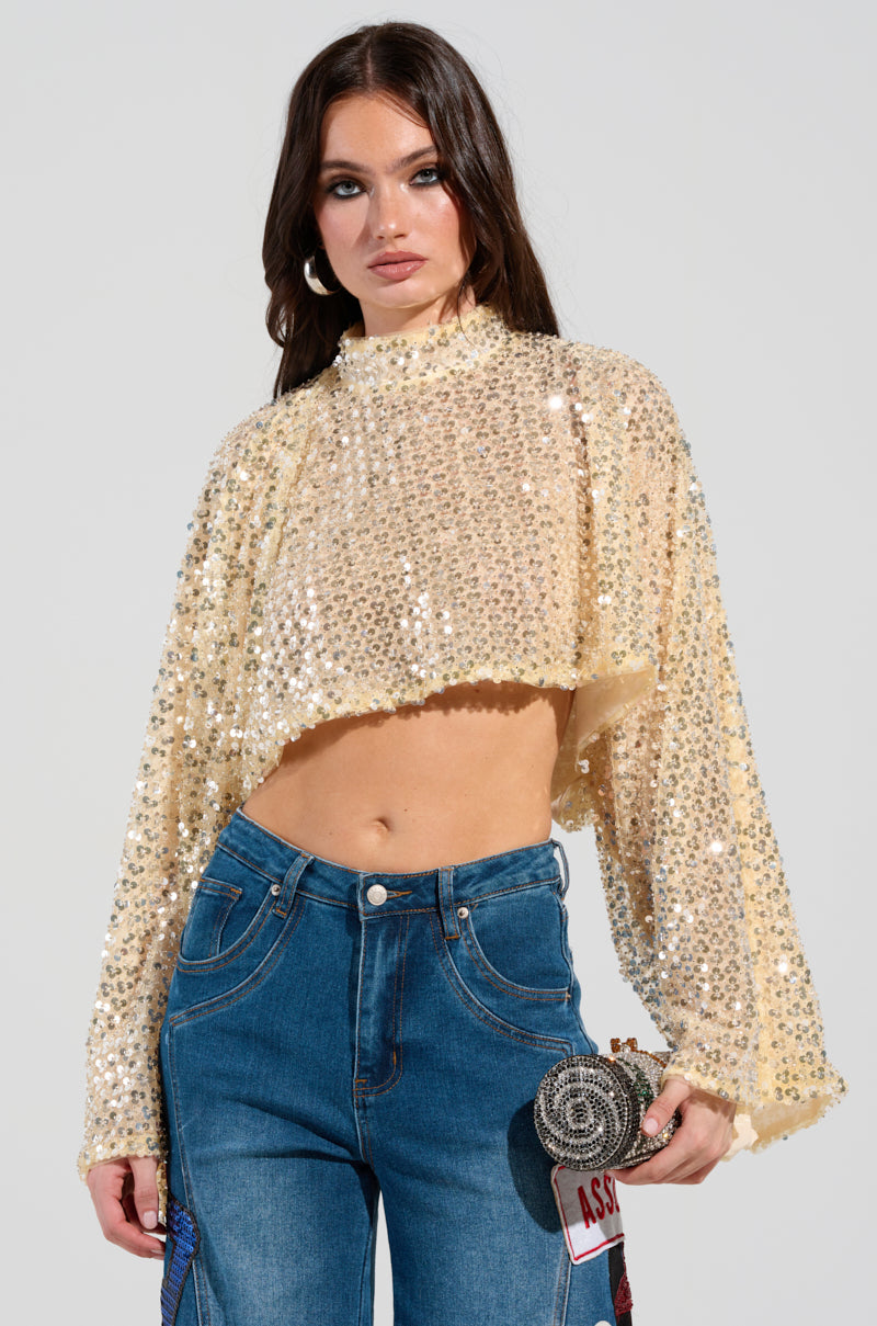 A woman models the SLYLINE SEQUIN BLOUSE, which features shimmering silver sequins and elegant long sleeves. She complements it with high-waisted blue jeans and carries a decorative clutch, crafting an ideal holiday party ensemble. Her long hair cascades down as she stands against a simple background.
