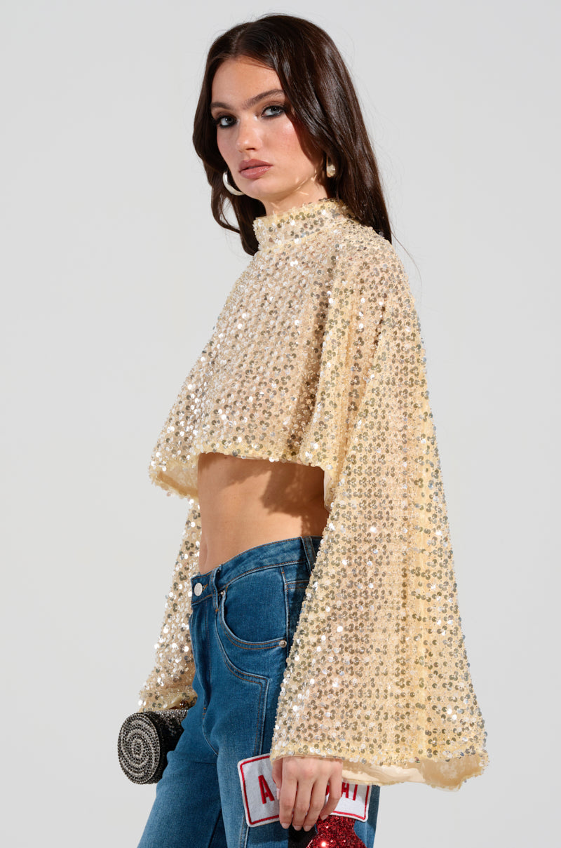 A woman wearing a dazzling SLYLINE SEQUIN BLOUSE stands in profile, capturing the ideal holiday party look. She complements it with blue jeans and holds a small round bag along with a clutch labeled "ASHISH." Her dark hair falls naturally against the plain light gray backdrop.