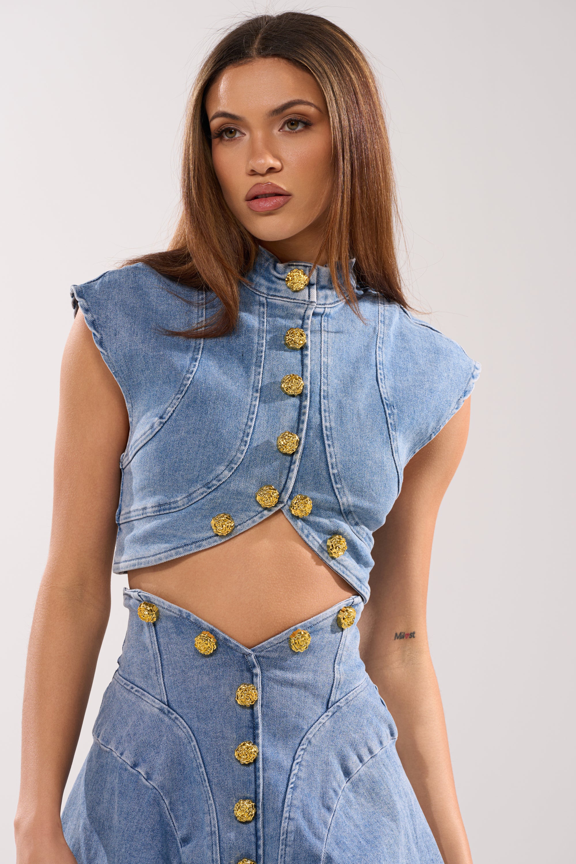 A woman wears the GIVE IT TO ME BABY DENIM BUTTON DOWN and matching high-waisted skirt with gold buttons, featuring a geometric waist cutout for a modern twist, against a plain background. 0