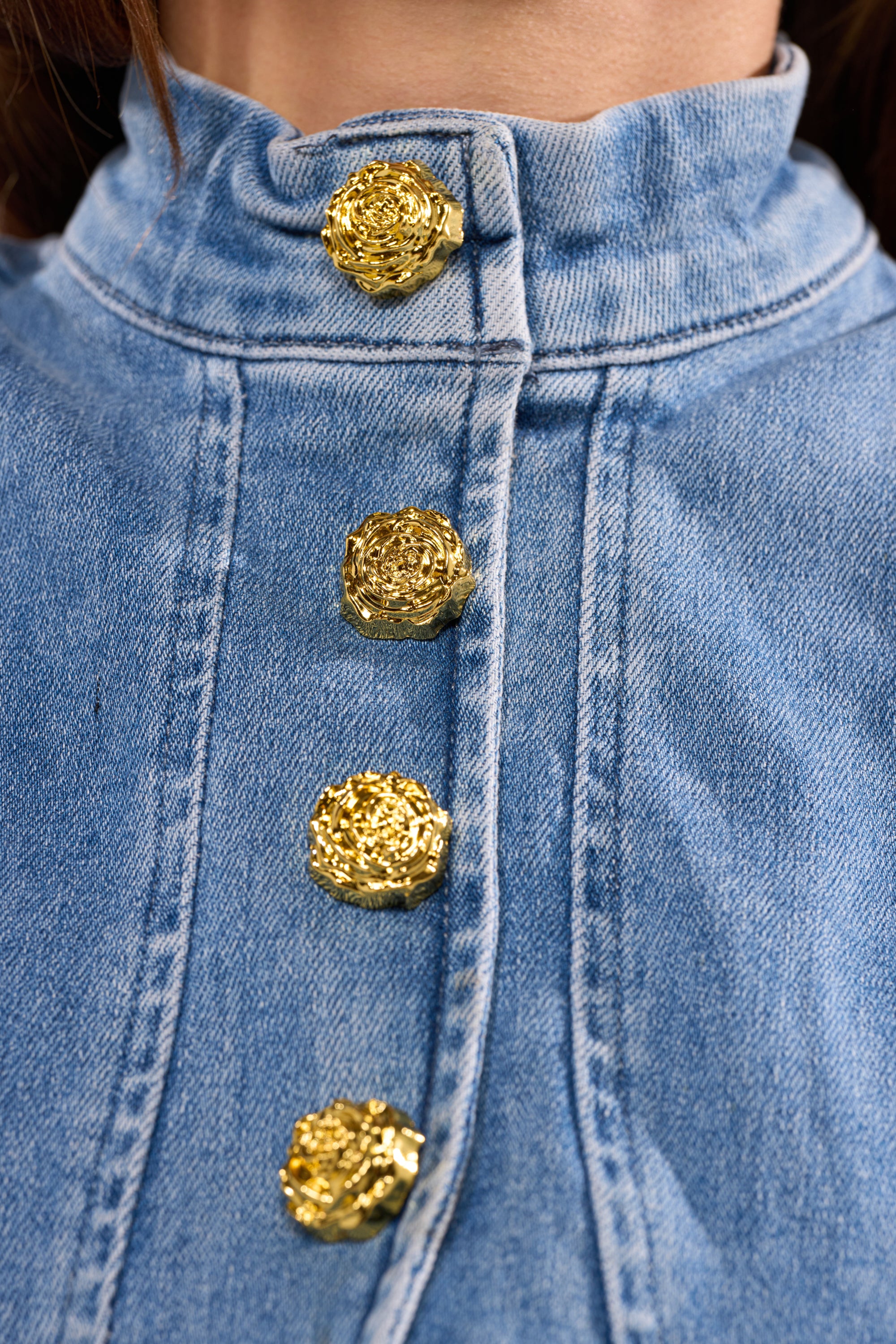 Close-up of the GIVE IT TO ME BABY DENIM BUTTON DOWN: a vintage-style blue denim top with a high collar and four ornate, textured gold buttons that stand out against the casual fabric. 10