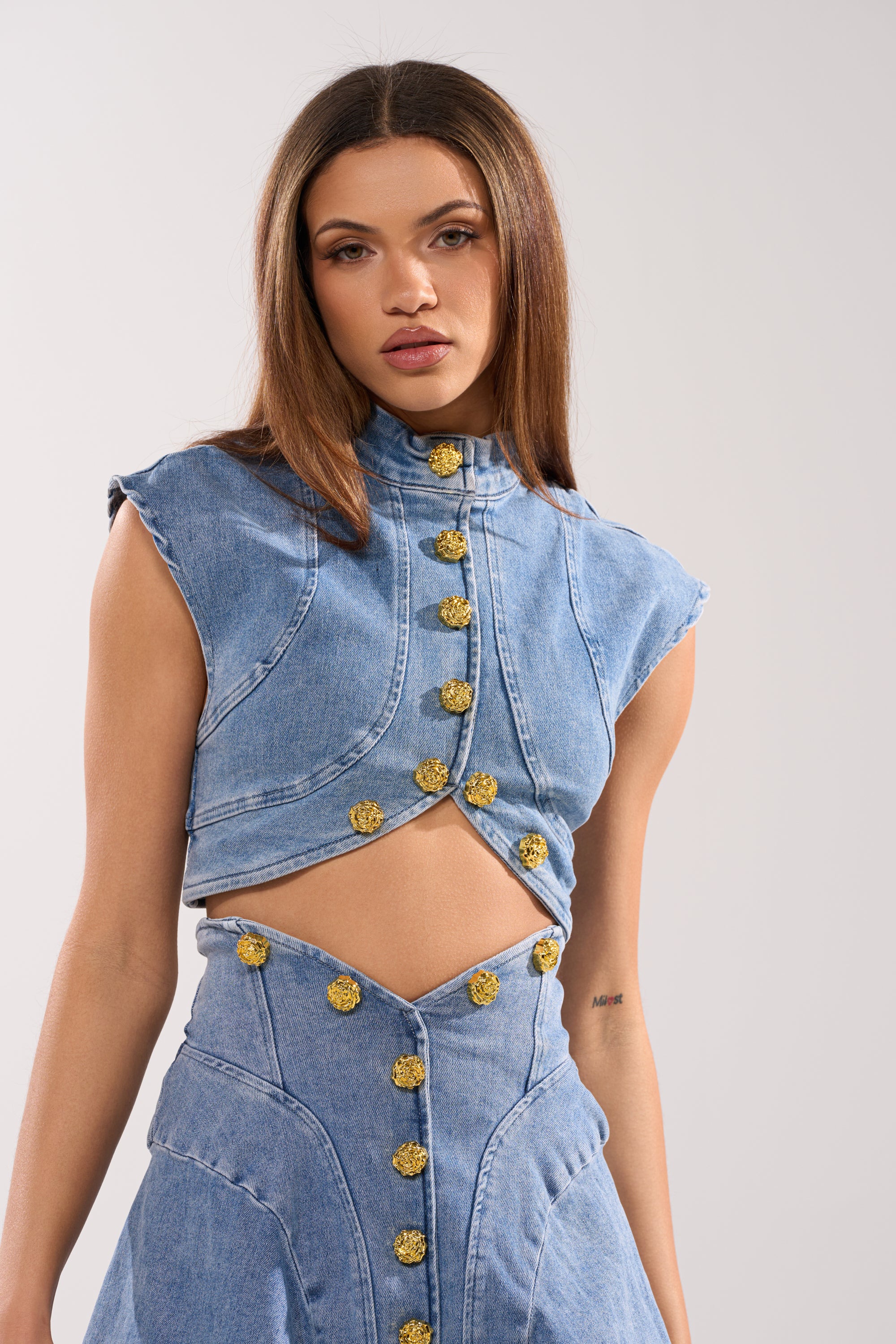 A woman with straight brown hair wears the GIVE IT TO ME BABY DENIM BUTTON DOWN, a vintage-inspired cropped top with gold buttons, paired with a matching high-waisted skirt, standing against a plain light background. 3