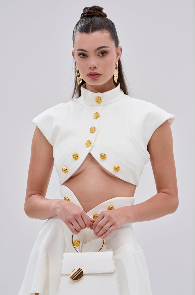 A woman wears the THINK ABOUT ME SLEEVELESS BLOUSE IN WHITE, a chic cropped top with metallic gold buttons, high collar, and cap sleeves, styled with a matching skirt. She accessorizes with a white handbag with gold handle and gold earrings. Her hair is pulled back. 0