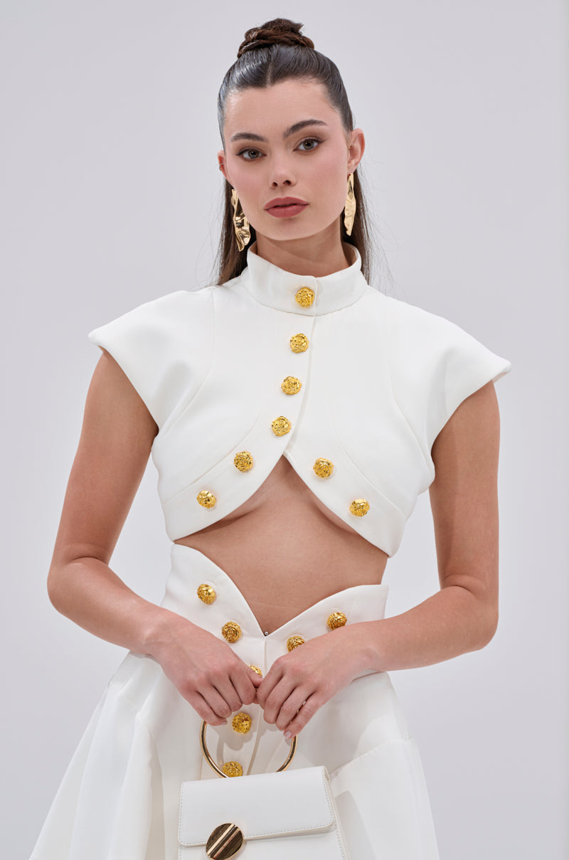 A woman wears the THINK ABOUT ME SLEEVELESS BLOUSE IN WHITE with metallic gold buttons and a matching high-waisted skirt, carrying a white handbag. She accessorizes with gold earrings, sleek makeup, and a high ponytail. 1