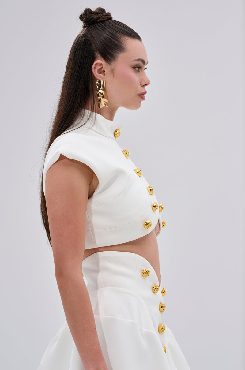 A woman with long, dark hair styled half-up wears the THINK ABOUT ME SLEEVELESS BLOUSE IN WHITE with a skirt featuring metallic gold buttons and large gold floral earrings, standing in profile against a plain light background. 2