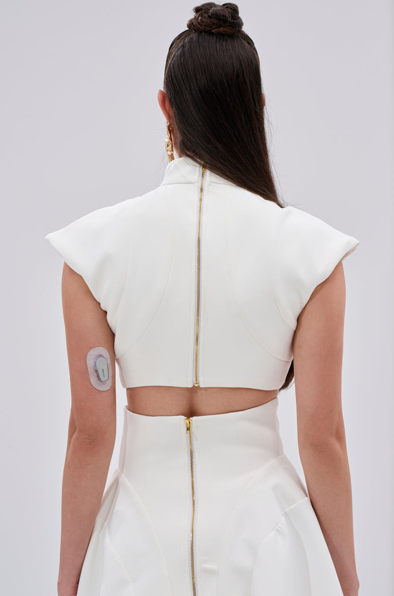 A person with long dark hair, seen from behind, wears the THINK ABOUT ME SLEEVELESS BLOUSE IN WHITE—structured and sleeveless with metallic gold buttons and an open back. A circular medical patch is on their upper left arm against a plain light background. 3