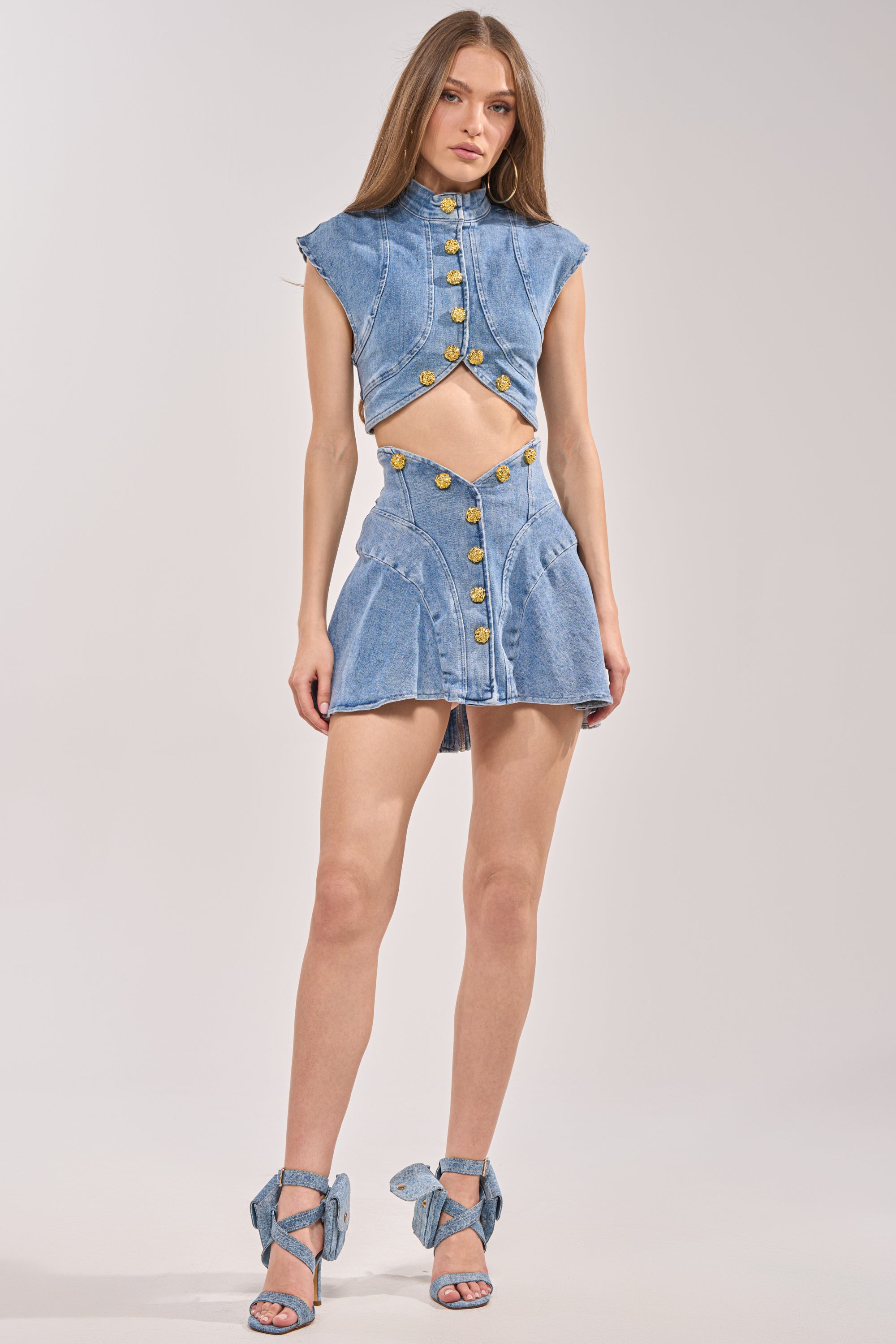 A woman stands against a plain light background wearing the GIVE IT TO ME BABY DENIM MINI SKIRT paired with a sleeveless, high-neck denim crop top with gold buttons and matching denim high-heeled sandals with bows. 6