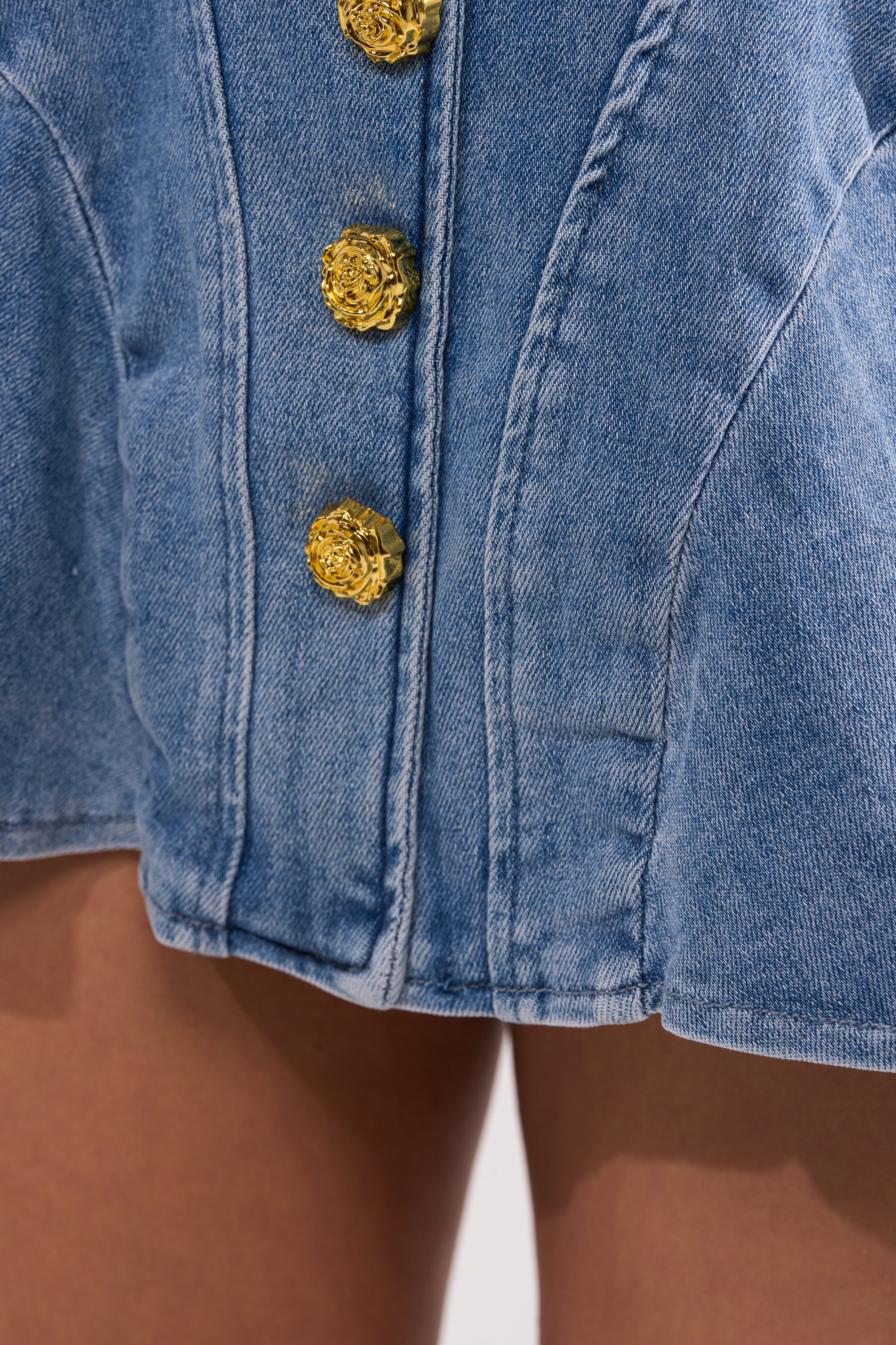 Close-up of the blue denim GIVE IT TO ME BABY DENIM MINI SKIRT featuring gold buttons. The high-waisted skirt flares slightly above bare legs. 7