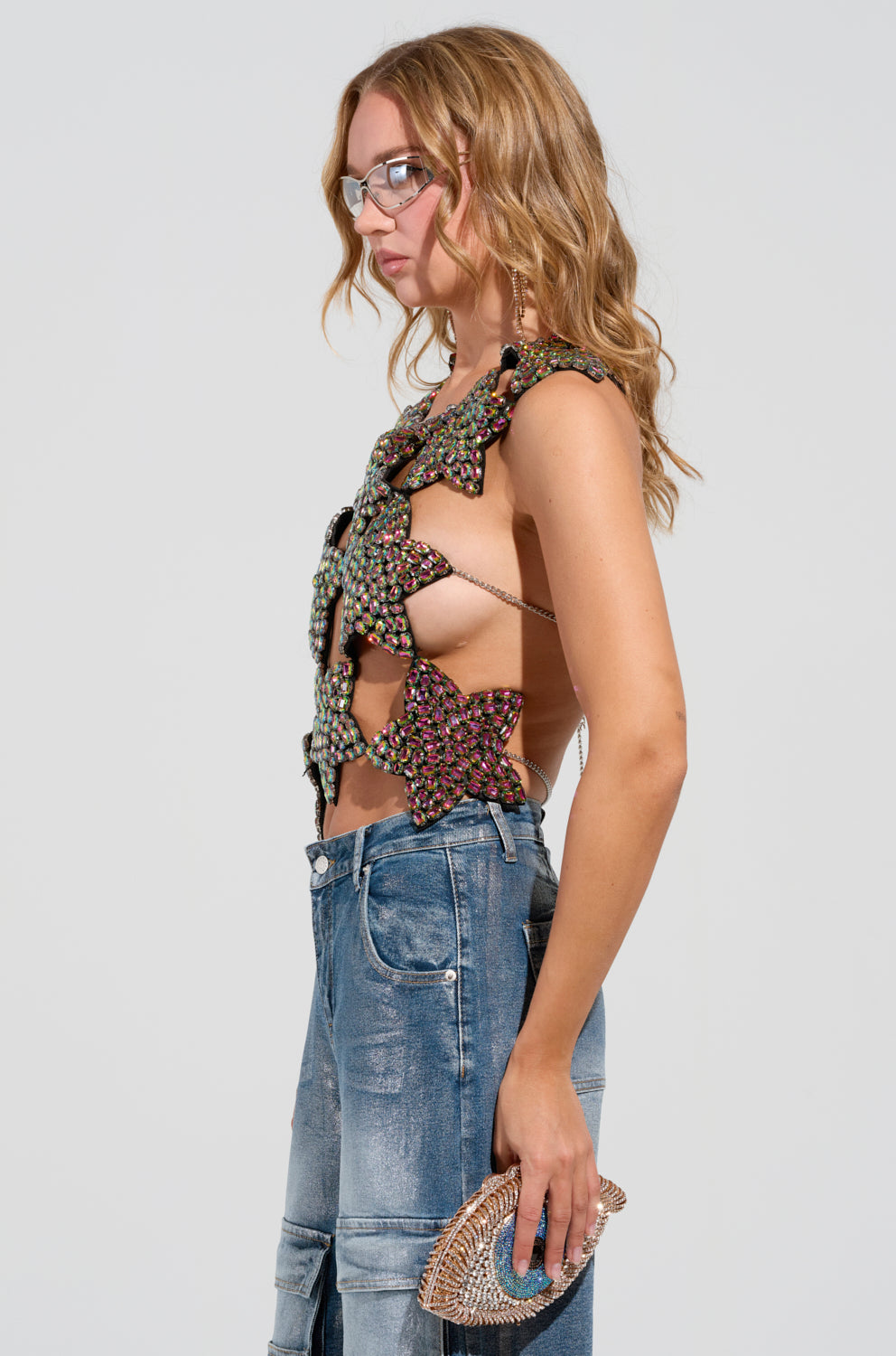 A woman with long hair wears the I'M THE STAR CUT OUT EMBELLISHED TOP paired with distressed jeans. She holds a small studded clutch with chain link straps, standing in profile against a plain white background.
