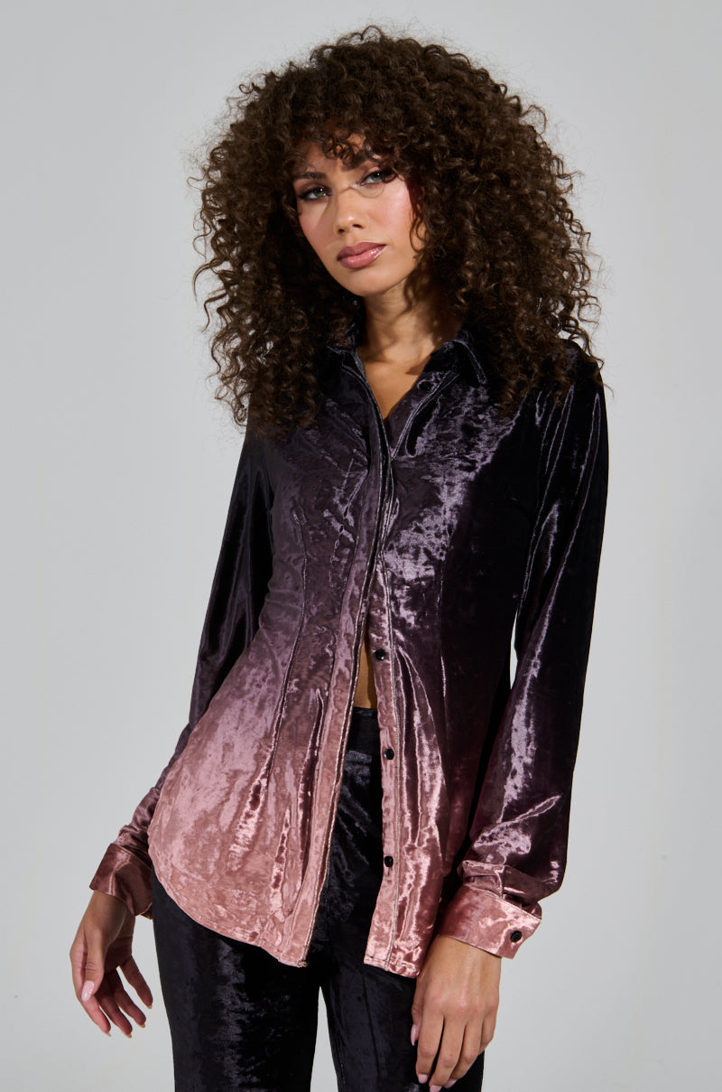 A person with curly hair is wearing the "NEVER SWITCH UP OMBRE BUTTON DOWN BLOUSE IN BLACK," crafted from crushed velvet and transitioning from pink to black, paired with matching pants. This holiday outfit is finished off with a collared shirt against a plain light gray background.