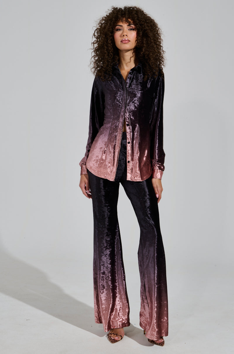 A person with curly hair models a chic holiday ensemble featuring the "Never Switch Up Ombre Button Down Blouse in Black," which transitions elegantly from black to mauve. The wide-legged pants match perfectly, creating a shiny texture as they pose gracefully against a plain background.
