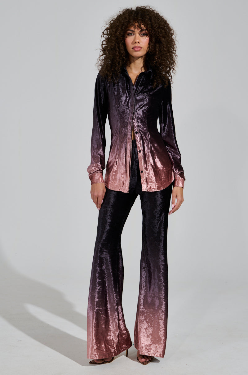 A person with curly hair stands wearing a holiday ensemble. The outfit features the "Never Switch Up Ombre Button Down Blouse in Black," which transitions from dark to light pink, paired with flared pants. They pose elegantly against a plain background.