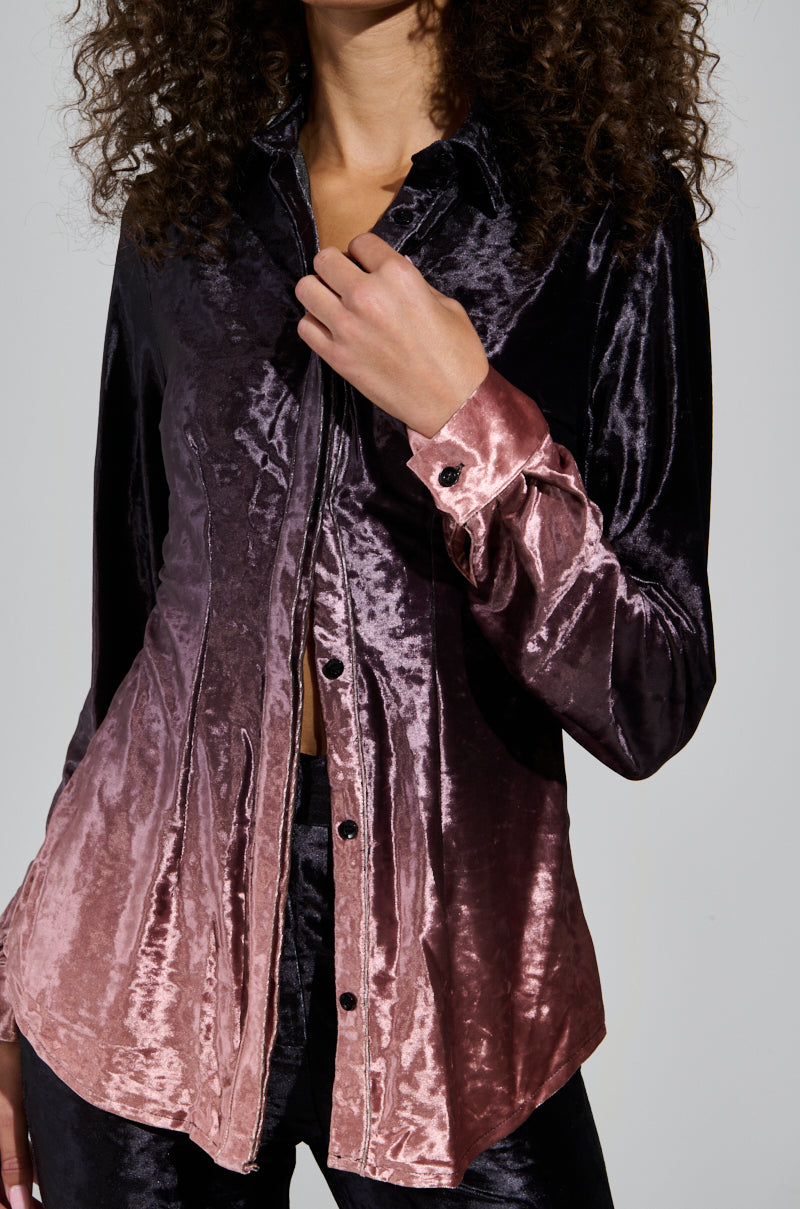 Clad in the NEVER SWITCH UP OMBRE BUTTON DOWN BLOUSE IN BLACK, a person showcases an ombre gradient from dark purple to pink, crafted from lustrous crushed velvet. With curly hair, they pose against a plain background, completing the ideal holiday ensemble.