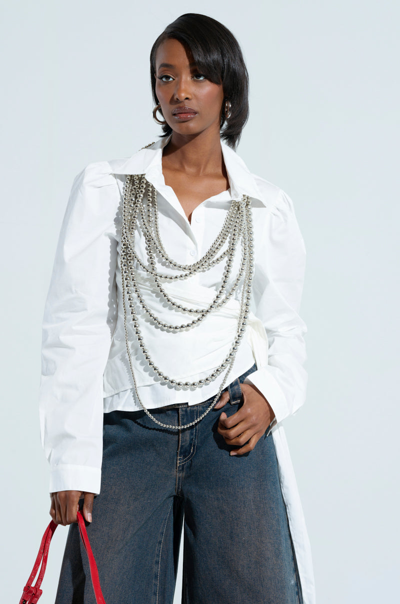 A person is wearing the BUILT TO LAST POPLIN BUTTON DOWN SHIRT paired with layered silver chains across the chest and wide-leg dark denim pants. They are holding a red bag while standing against a plain light background.
