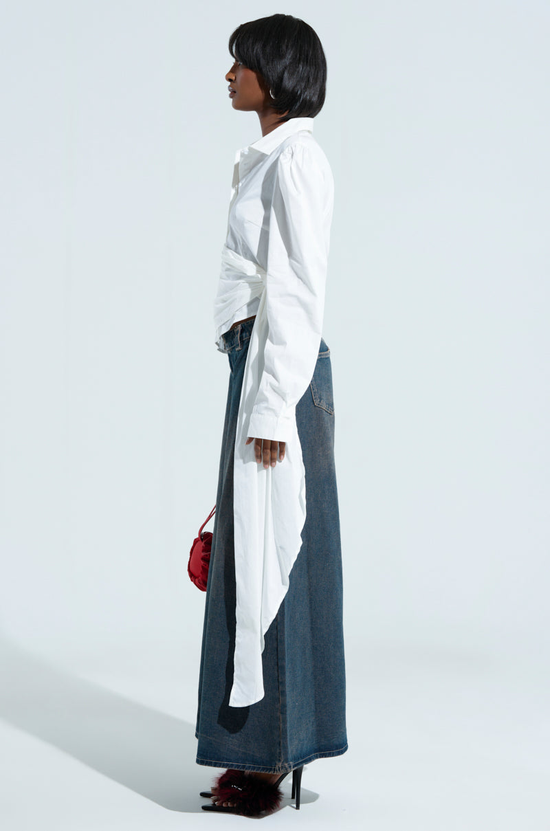 A person stands in profile against a light background, wearing the "BUILT TO LAST POPLIN BUTTON DOWN SHIRT" with asymmetrical tails, a long denim skirt, and high-heeled shoes. They hold a small red bag.