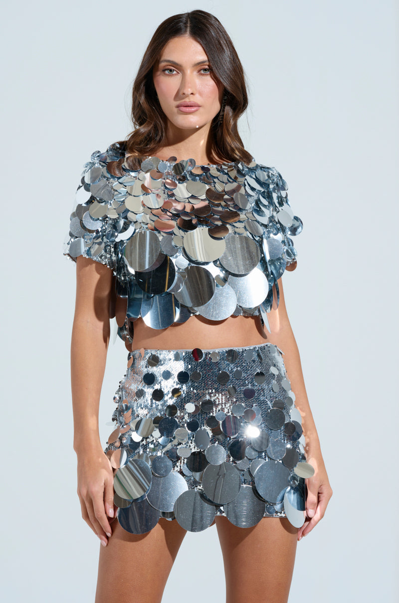 Wearing a captivating COUNTDOWN TO MIDNIGHT TOP, adorned with dazzling reflective discs and sequins, a person poses against a neutral background. The cropped top and skirt ensemble complements their confident expression and long brown hair, creating an enchanting look.