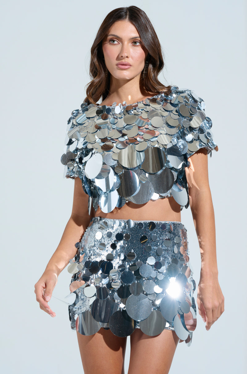 Set against a light blue backdrop, a person strikes a pose in the shimmering COUNTDOWN TO MIDNIGHT TOP, adorned with large silver sequins. The cropped design reflects light, creating an alluring effect.