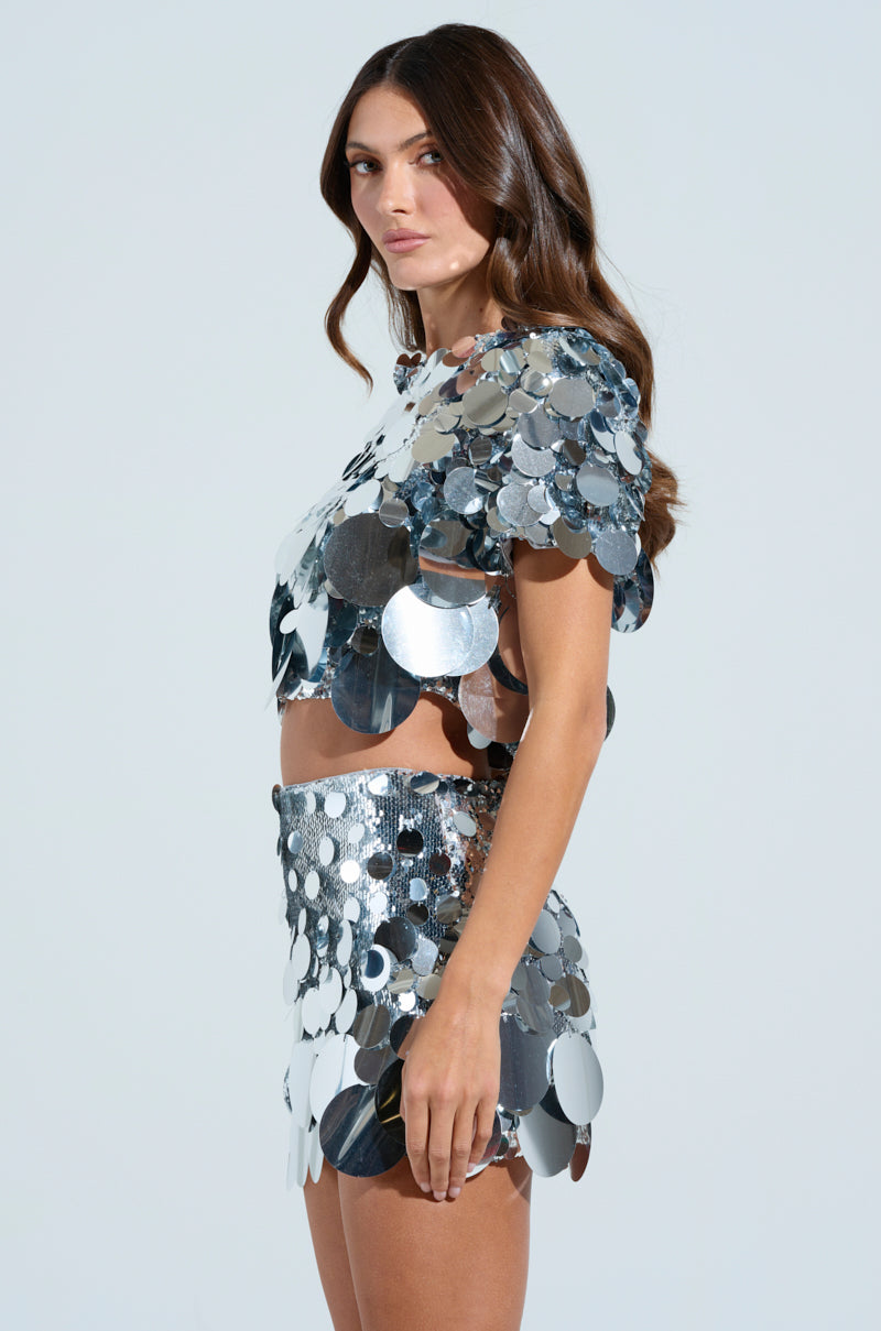 A woman in the COUNTDOWN TO MIDNIGHT TOP and matching skirt with large silver sequin discs stands against a plain background, her long wavy brown hair styled loose, perfectly capturing the essence of a countdown to midnight celebration.