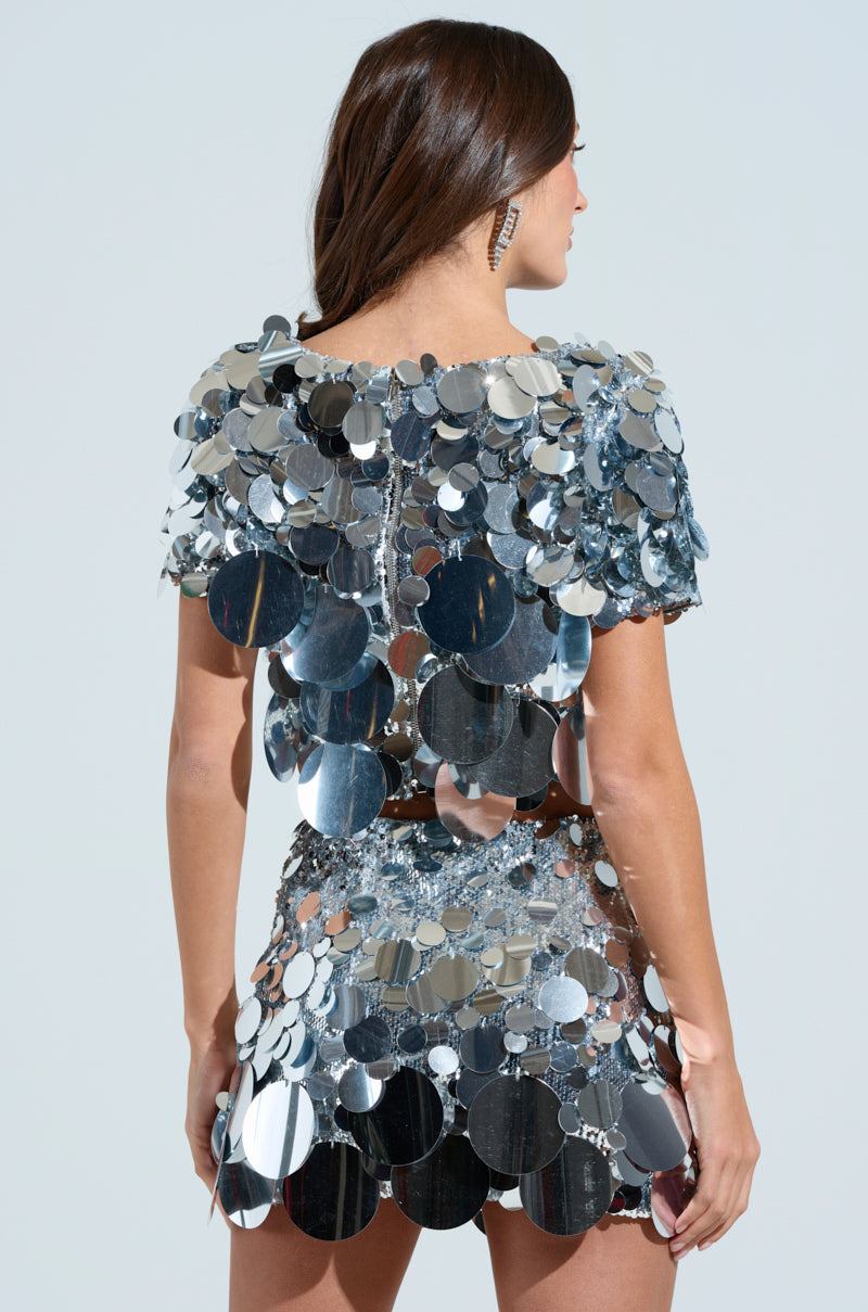 A woman with long brown hair stands sideways, wearing the shiny COUNTDOWN TO MIDNIGHT TOP adorned with large silver sequined discs that reflect light. The top has short sleeves and a short hemline, creating a futuristic look against a plain background.