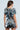 A woman with long brown hair stands sideways, wearing the shiny COUNTDOWN TO MIDNIGHT TOP adorned with large silver sequined discs that reflect light. The top has short sleeves and a short hemline, creating a futuristic look against a plain background.