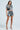 A woman confidently wears the COUNTDOWN TO MIDNIGHT top with a mini skirt, both adorned in silver sequins and large circular embellishments. Her matching strappy heels add glamour against the simple light gray background as she preps for the Countdown to Midnight.