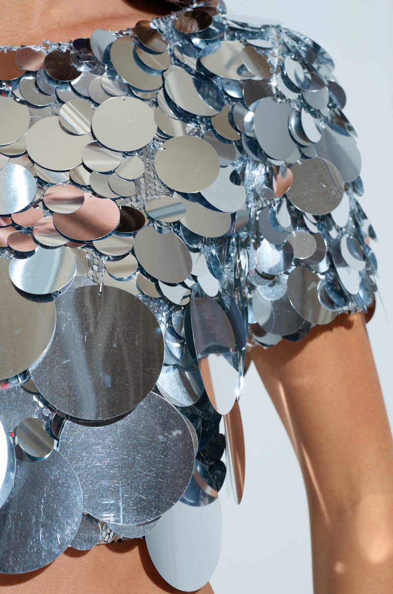 A close-up shows a person wearing the COUNTDOWN TO MIDNIGHT TOP, featuring large reflective silver sequins. These shiny discs gleam in the light, giving a metallic and futuristic look. Only the shoulder and upper torso are visible against a neutral background.