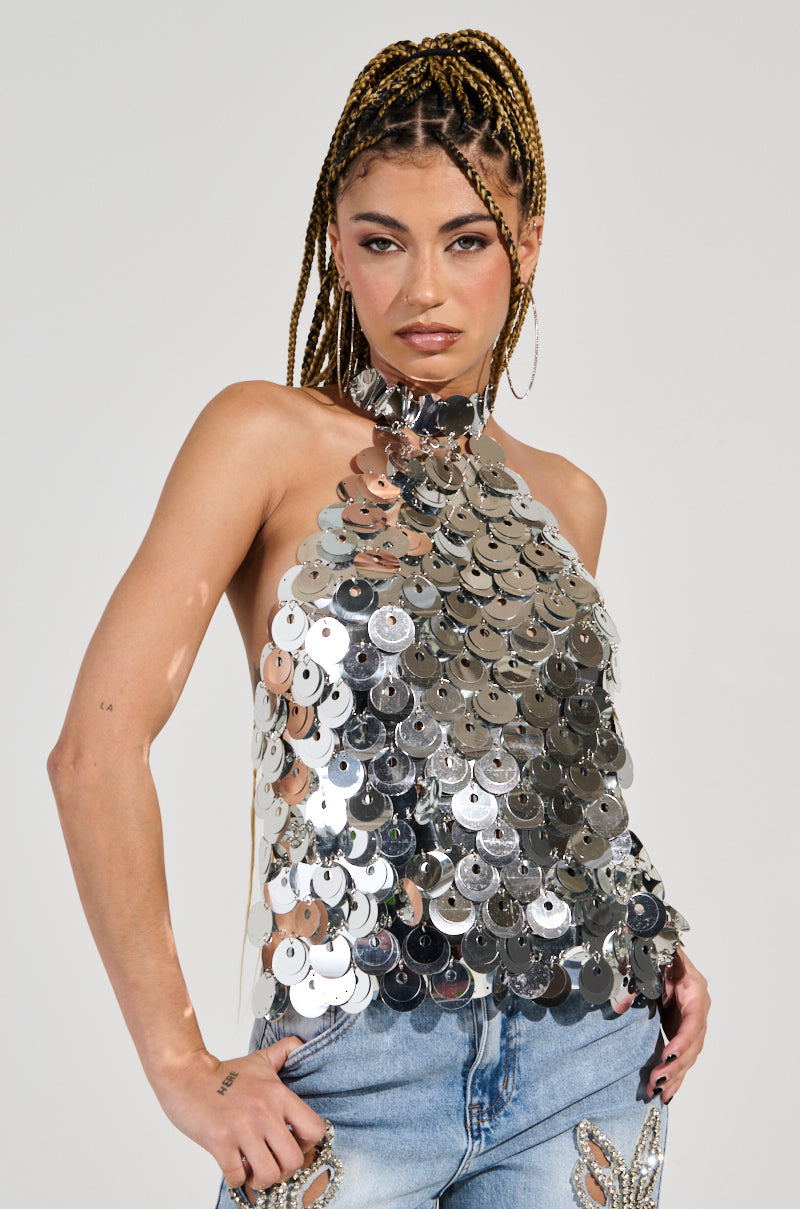 A person with braided hair wears the PARTY ALL NIGHT SEQUIN HALTER NECK TOP, adorned with large silver sequin discs, paired with light blue jeans featuring beaded embellishments. They stand confidently against a plain background, looking directly at the camera. 2