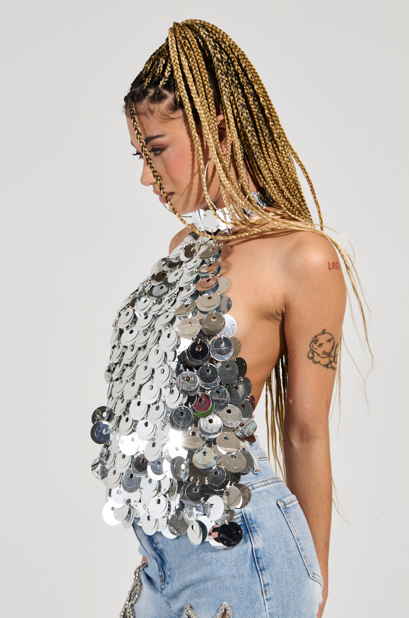 A woman with long braids is shown in profile, wearing the PARTY ALL NIGHT Sequin Halter Neck Top adorned with shimmering silver sequin discs and light blue jeans. She has a tattoo of a cherub on her arm against a plain white background. 3