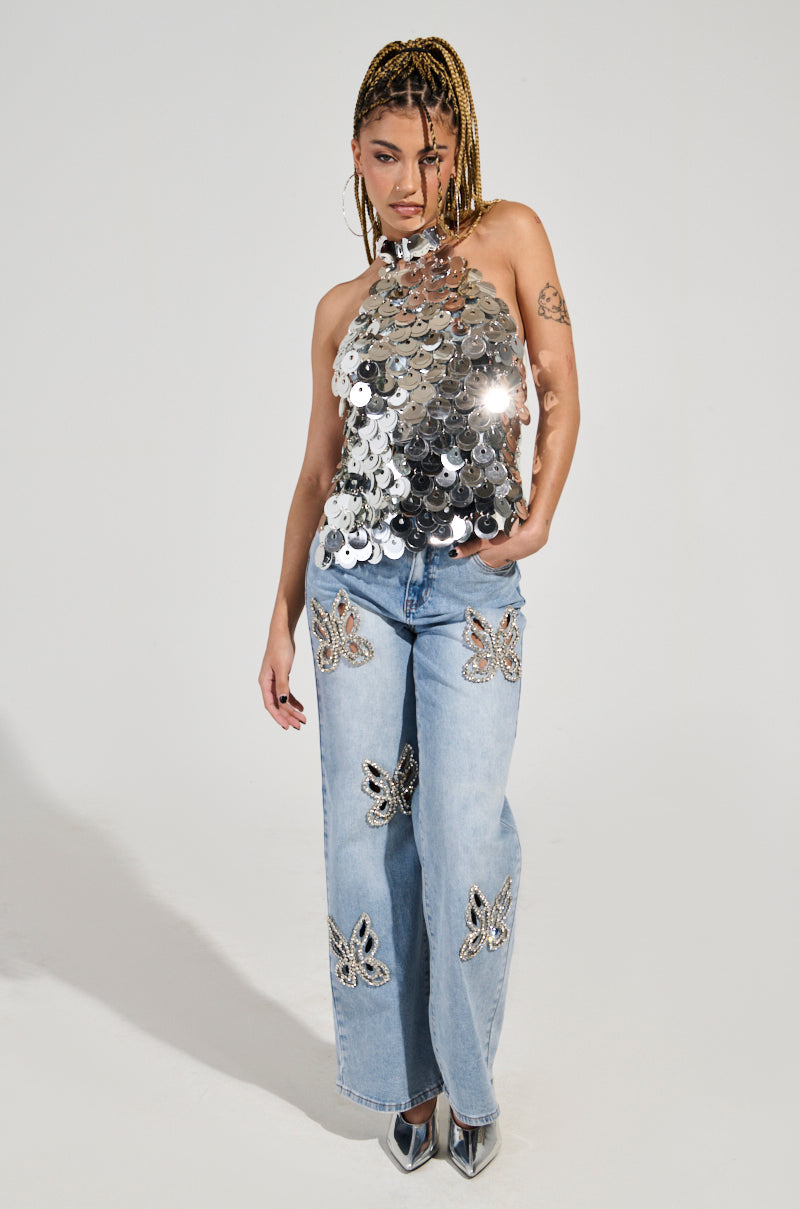 A person poses against a plain background wearing the PARTY ALL NIGHT SEQUIN HALTER NECK TOP, featuring shiny silver sequin discs. They sport light blue jeans decorated with sequin butterfly designs and have braided hair paired with metallic pointed shoes. One arm is slightly bent while the other remains relaxed. 5