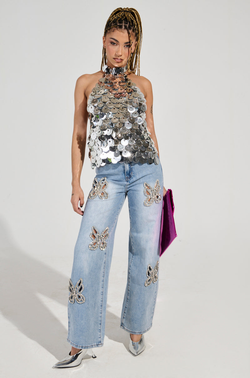 A person wearing the PARTY ALL NIGHT SEQUIN HALTER NECK TOP with silver sequin discs and light blue jeans featuring butterfly embellishments. They hold a purple clutch and wear silver pointed heels, standing against a plain white background. 6