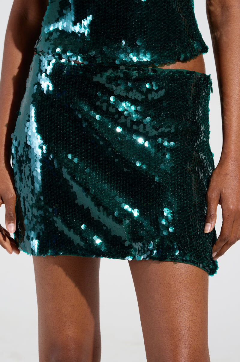 Wearing the "BEST IN THE CITY MINI SKIRT" by AKIRA Label, a person stands out with matching attire as the dark green sequins shimmer brilliantly against a plain background. Their hands rest at their sides, effortlessly capturing the light's sparkle.