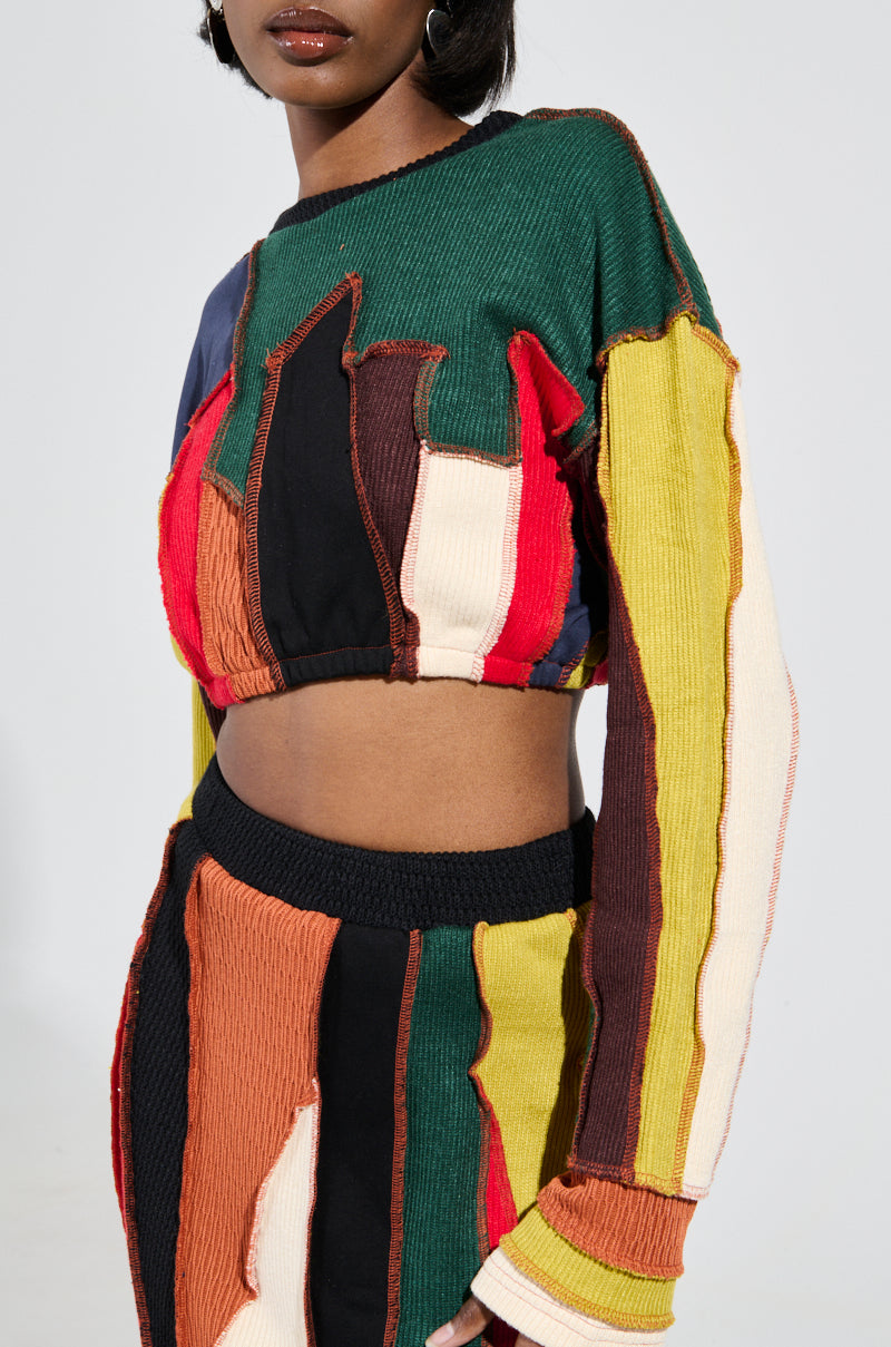 A person wearing the BEAST MODE SWEATER, a textured long-sleeve crop top with a matching skirt, both featuring a chic cropped silhouette. The ensemble displays vertical stripes in vibrant shades of green, yellow, red, black, and brown against a plain and neutral background. 0
