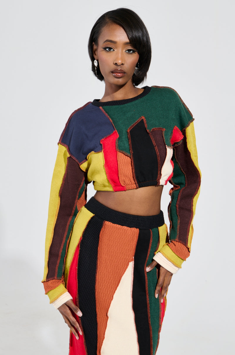 A woman poses confidently in the vibrant BEAST MODE SWEATER, a colorful two-piece outfit with bold patchwork construction. The ensemble includes a long-sleeved top and knee-length skirt, highlighting striking hues such as green, red, yellow, black, and brown. Her shoulder-length hair and hoop earrings enhance her fierce vibe. 1