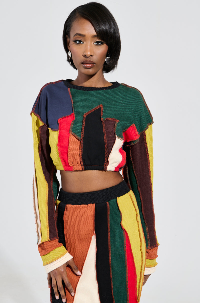 A person showcases the BEAST MODE SWEATER, featuring a vibrant patchwork crop top and skirt set against a plain background. This eye-catching ensemble stands out with its cropped silhouette and vertical stripes in red, yellow, green, and black. The person's expression remains neutral. 2