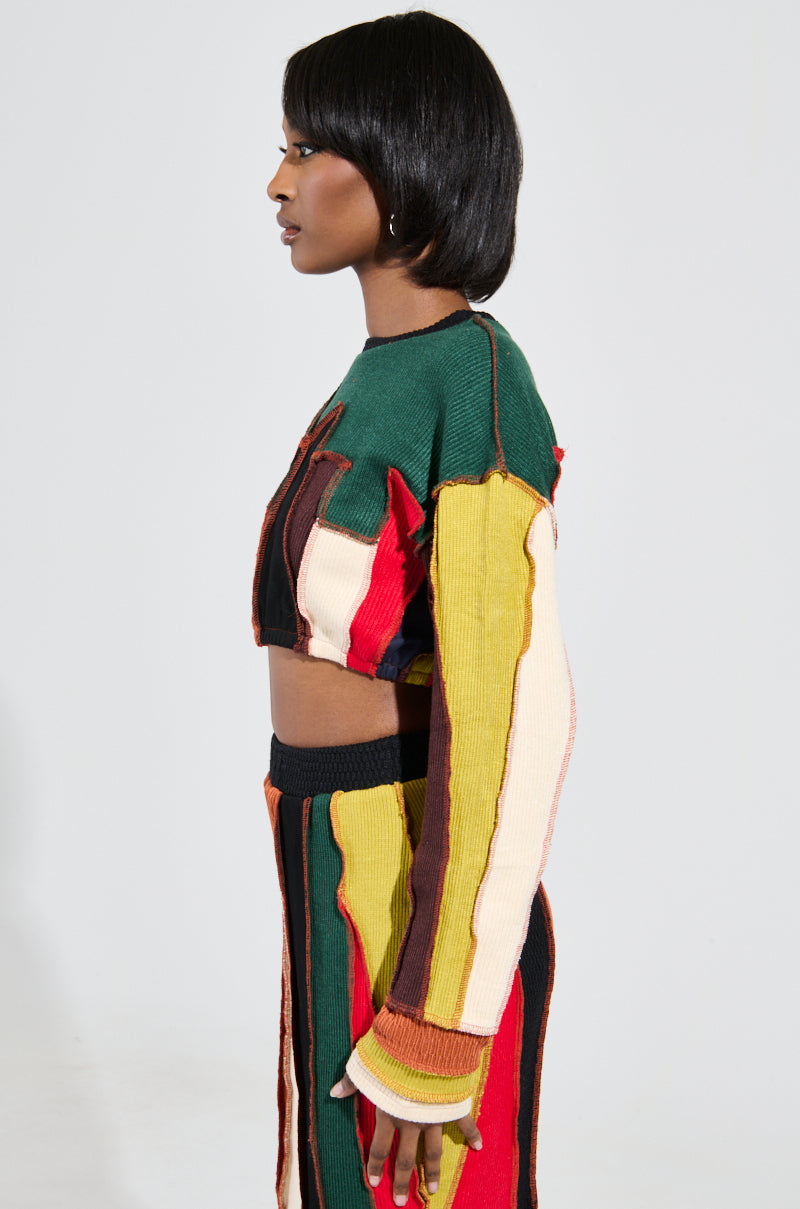 Dressed in the eye-catching BEAST MODE SWEATER, a person stands sideways showcasing a lively cropped outfit with vertical stripes in shades of green, yellow, red, and brown. Complemented by high-waisted pants against a simple white background, the long-sleeved top epitomizes patchwork construction flair. 3